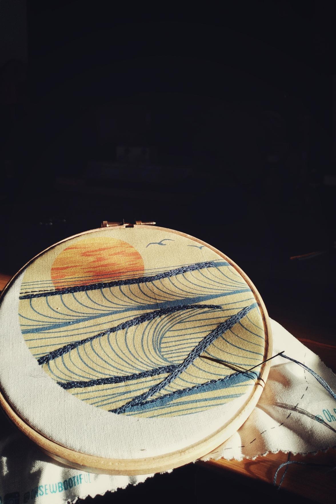 A vertical colour photograph of a pre-printed embroidery work. The design is a stylised sunset over the sea. Some of the needlework has been started. The hoop is on a strong beam of sun. The background fades into dark.