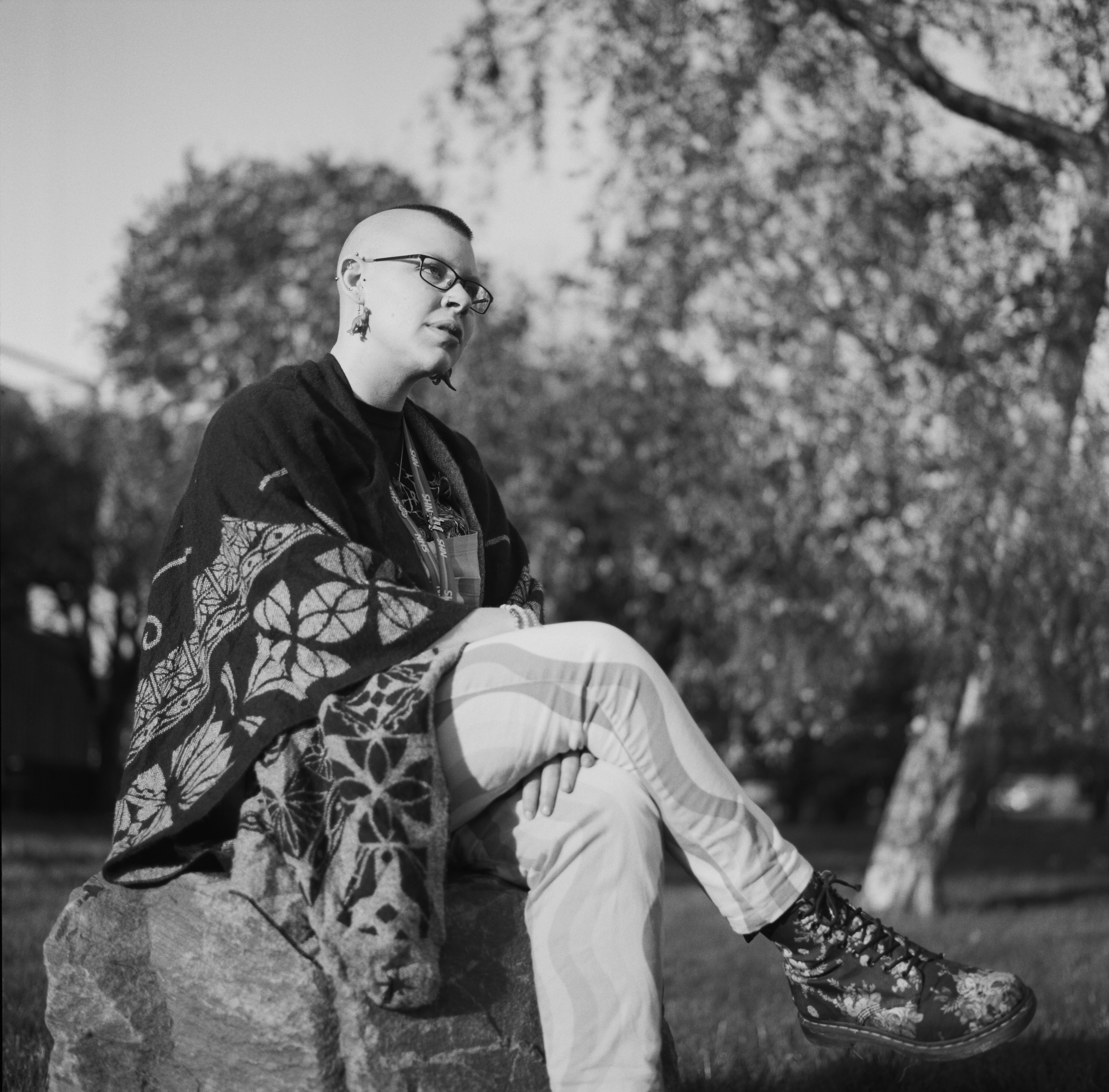 A black and white photography of a person sitting on a rock. They have short cropped hair and are wearing a long wollen top and rainbow trousers. They are looking off in the distance.