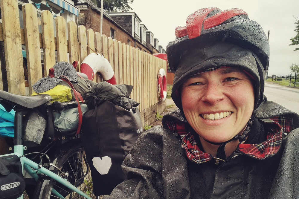 A horizontal colour photograph of me (a white person) smiling. I am wearing a dark green canva poncho and a red helmet. I am crouched by my bicycle. Only the rear of it is visible with panniers and bags loaded on it. The bicycle rests against a wooden picket fence. It is raining and everything, including me, is covered in drops of rain.