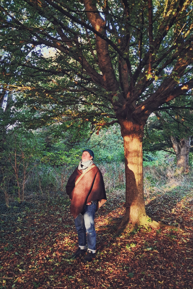 A vertical colour photograph of me standing by a tree in a park and bathing in the low lying golden rays of sunlight. I am a white person wearing a teal woolen beanie, a brown woolen cape, dark blue jeans, and black trainers.