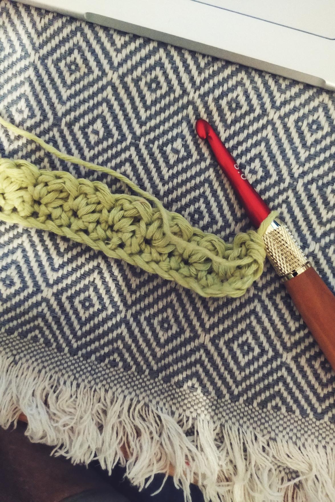 A vertical colour photograph of the corner of a light green crochet cloth. It's just the first rows of it. A crochet hook is attached to it. It has a wooden handle and a metallic red hook. The work is on a diamond patterned blue and white cloth. At the top of the image is the bottom of a laptop trackpad. At the bottom of the image is the blue and white cloth fringe (all white) and the little bit of my jumper (mostly in shadows).