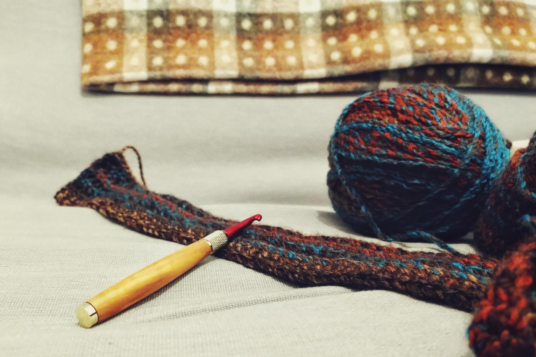 A horizontal colour photograph of a crochet scarf in progress laid on a grey sofa. The scarf is laid down with the crochet over it. On the right hand side of the image, the scarf starts to bundle on itself. The ball of yarn used is behind the scarf. In the background, a green and brown throw rests against the back of the sofa.