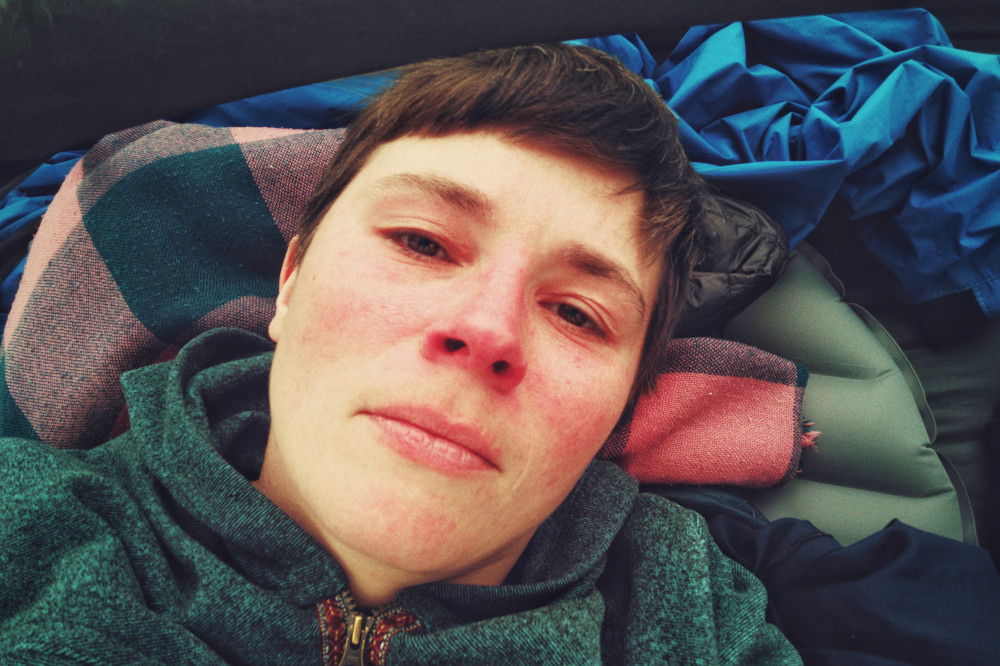 A horizontal colour photograph of my face lying on a sleeping mat covered by a blanket that acts as a pillow. I am wearing a green fleece. My face is red, my eyes watery from tears, my expression blank. I have short brown hair.