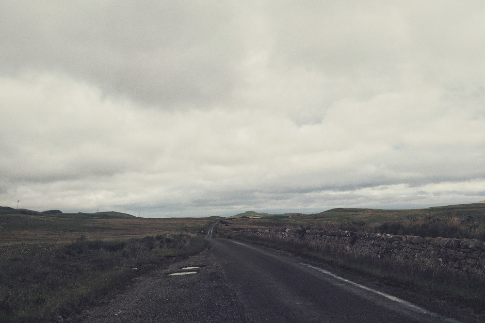 A horizontal colour photograph of a snaking single lane road disappearing into the distance. On either side is grass. One side is hemmed by a stone wall. In the distance, rounded hills are visible. The sky is under a layer of whitish grey clouds.