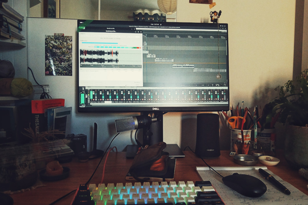 A horizontal colour photograph of a desk with a white, pink, and black mechanical keyboard. Around it is a pen pouch, a notepad with notes and a pen on it. The computer screen shows an audio editing software exporting audio. All around are plants, notebooks, some yarn, etc.
