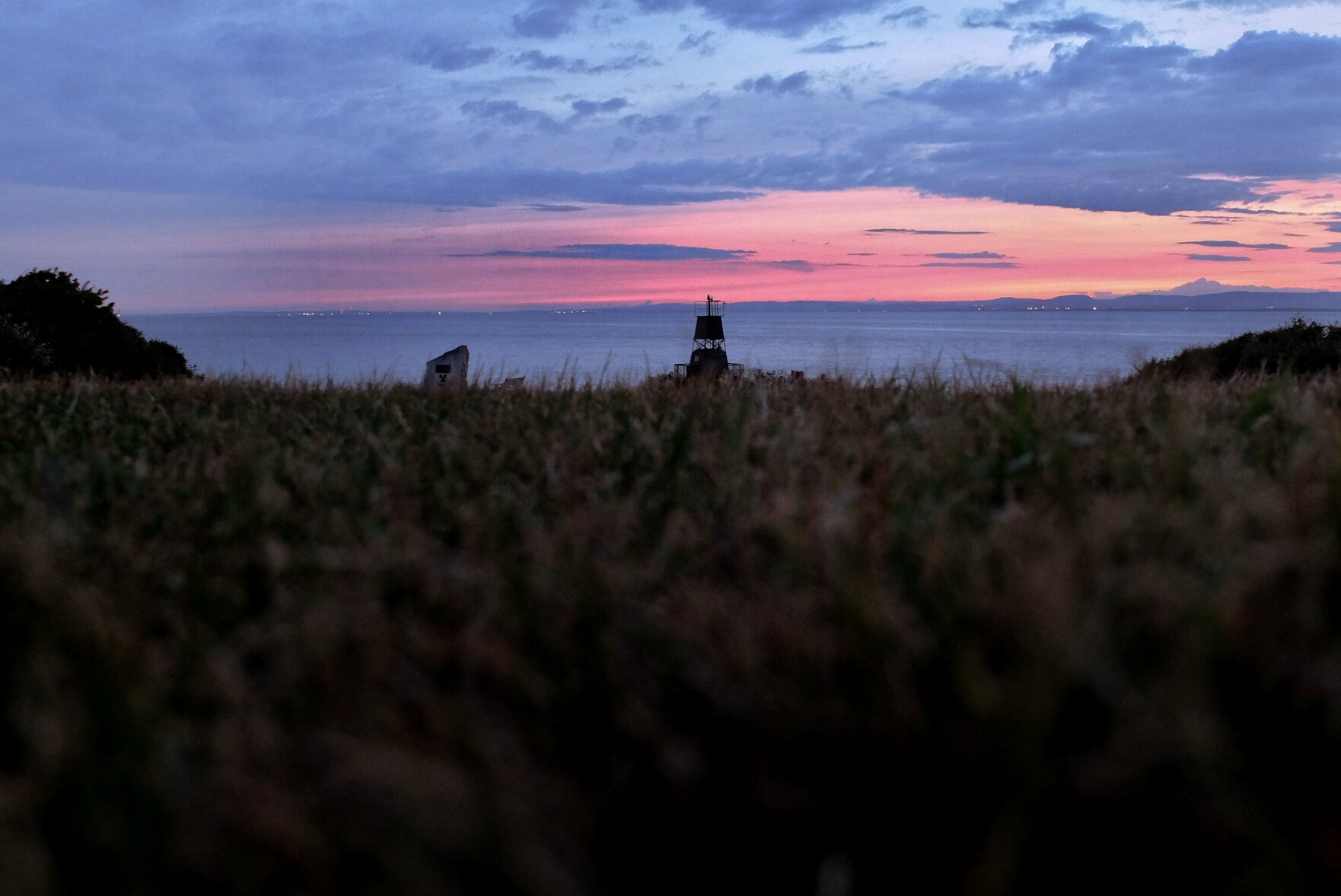 A horizontal colour photograph of the sunset over water taken from low down in the grass. The bottom two third of the image are taken by the grass. In the middle, the in the distance is a small dark lighthouse.  The water is still. There are clouds in the sky.