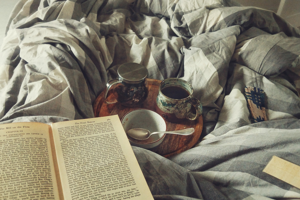 A horizontal colour photograph of a grey bed duvet with on top of it an open book, a couple of bookmarks, and a tray with a tea pot, mug, and small empty bowl with a spoon in it.