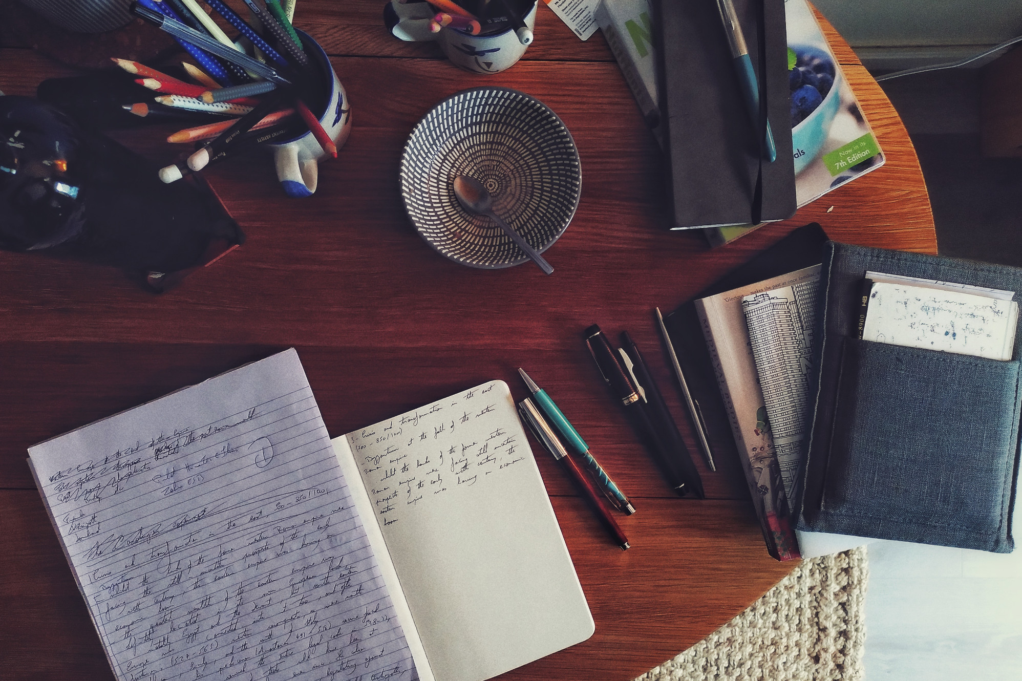 A horizontal colour photograph of a wooden table covered with a notebook and a notepad full of scribbled notes. Pens and books lay next to those. All around are various objects such as an empty bowl, pencils in small mugs.