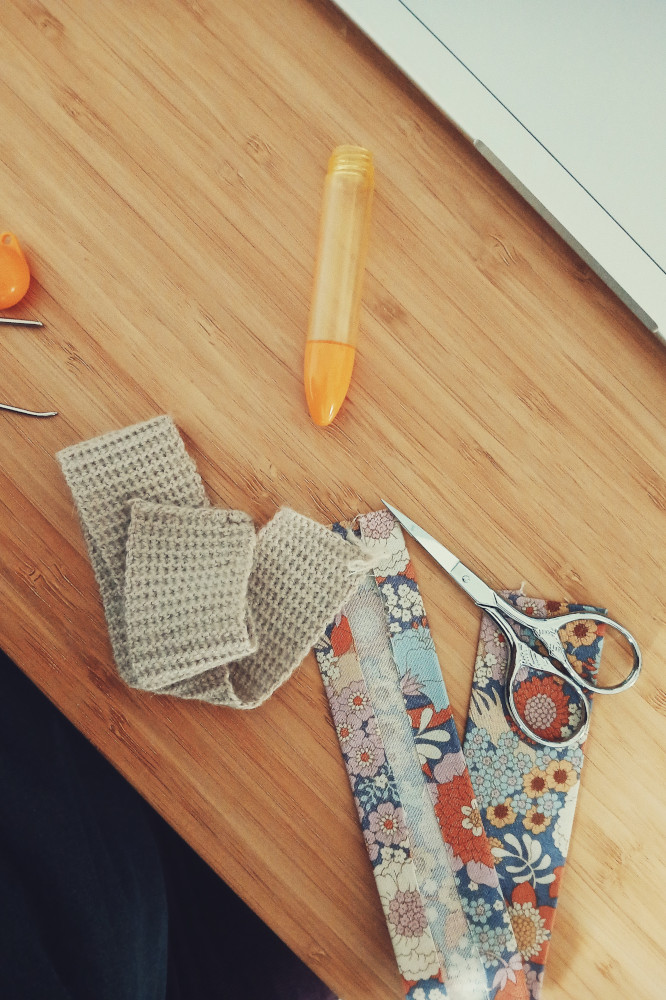 A vertical colour photograph of a tangled strip of crochet fabric next to a cut up lining. Around them are a pair of small scissors, a case for tapestry needles, and some tapestry needles cut up by the frame.