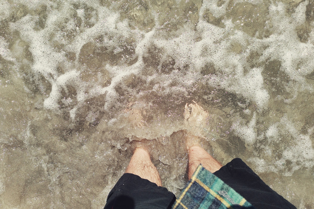 A horizontal colour photograph of my feed in foamy water. My legs are bare and hairy. I am wearing black shorts. A corner of my green and yellow checked shirt flies in the frame.