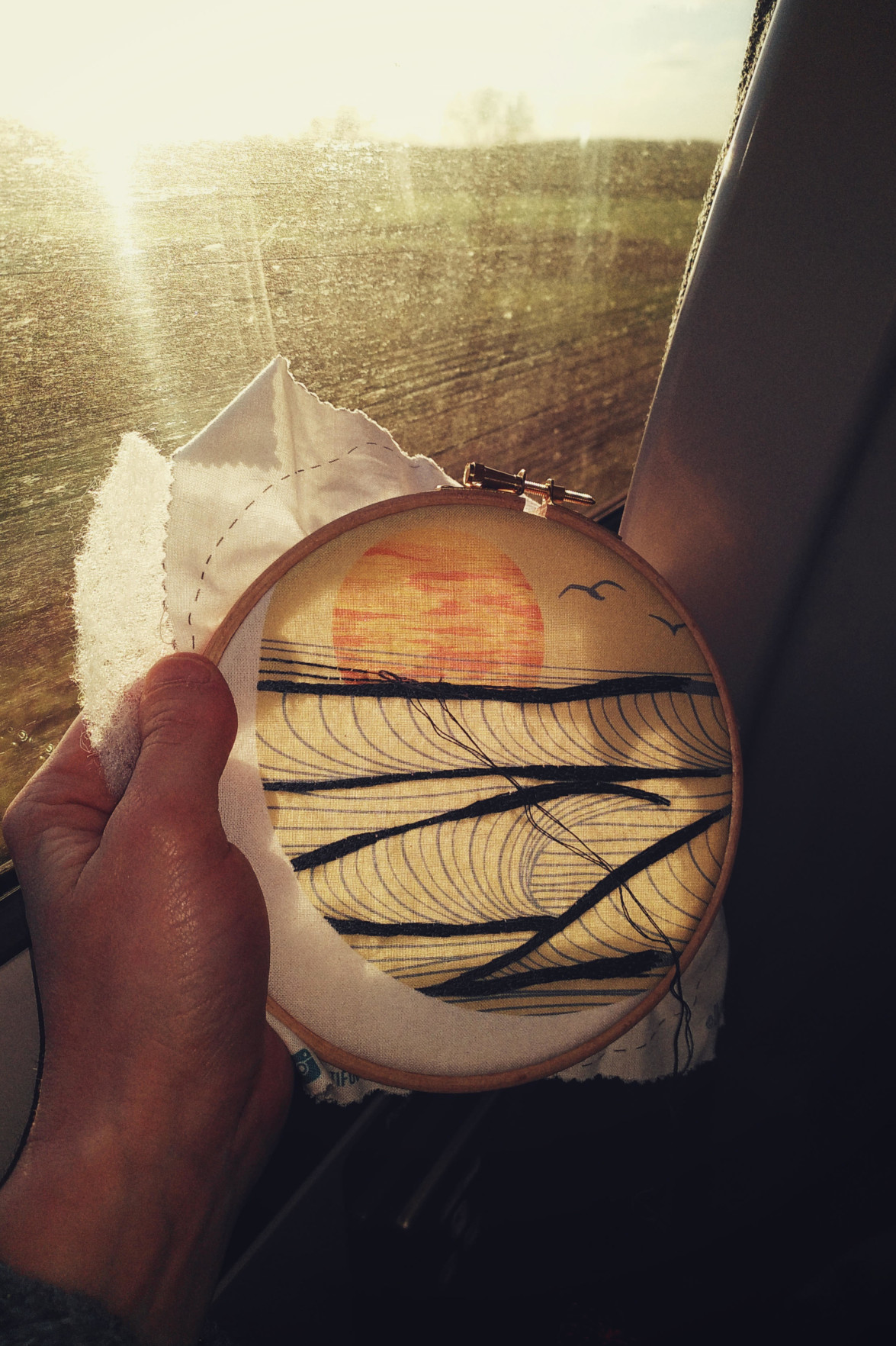 A vertical colour photograph of an embroidery hoop with a stylised sunset printed on the cloth. Embroidery is in progress. I am holding the hoop by a train window bathed in light.